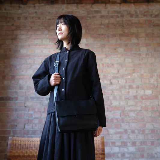 Person dressed in black with black leather shoulder bag standing in front of brick wall