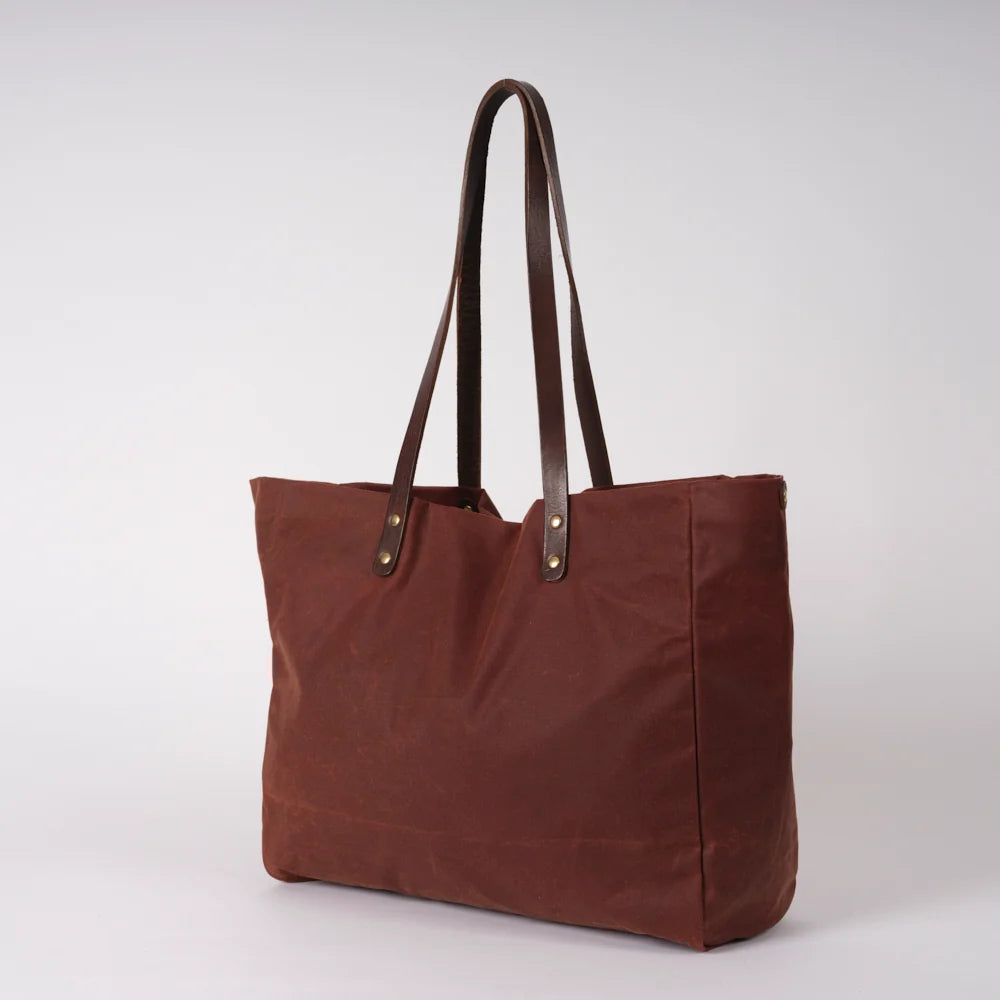 A brown canvas tote bag with dark brown leather shoulder straps and brass-tone rivets, photographed on a clean light gray studio background.