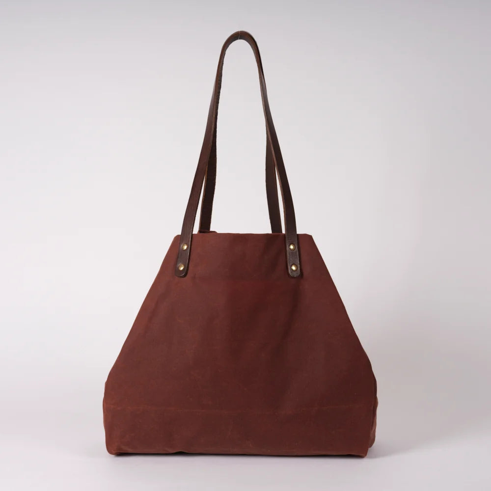 A brown canvas tote bag with dark brown leather shoulder straps and brass-tone rivets, photographed against a neutral light gray studio background.