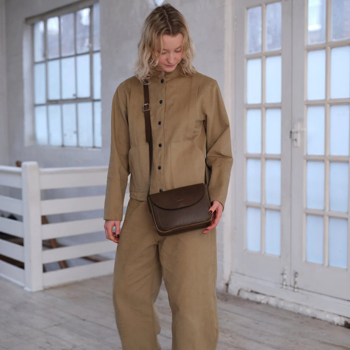 Woman dressed in sand colored jacket and pants with sage green leather crossbody bag with a curved flap and a long adjustable strap, photographed on a light gray studio background.