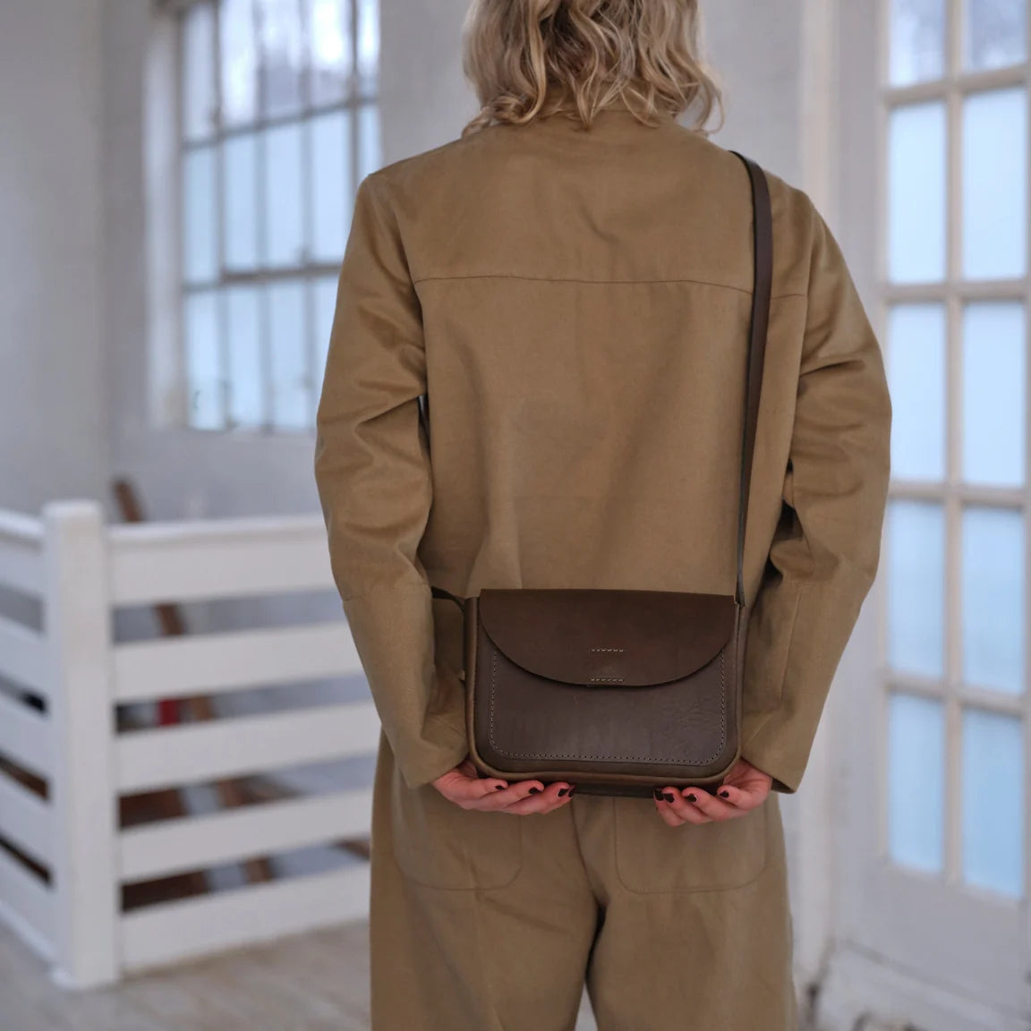 Rear view of woman dressed in sand colored jacket and pants with sage green leather crossbody bag with a curved flap and a long adjustable strap, photographed on a light gray studio background.