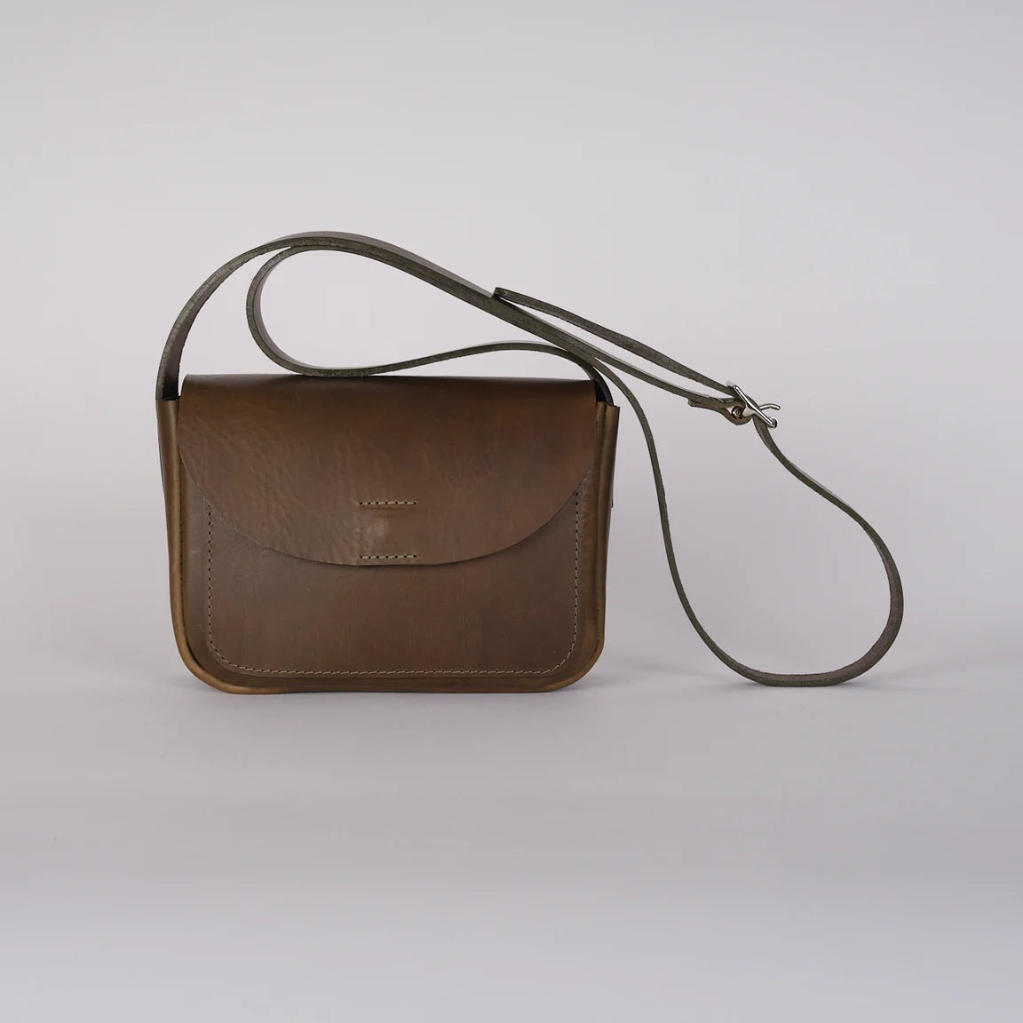 A sage green leather crossbody bag with a curved flap and a long adjustable strap, photographed on a light gray studio background.