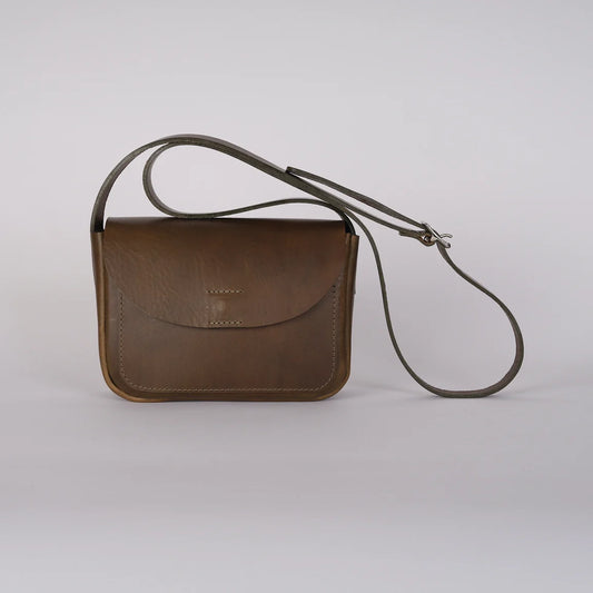 A sage green leather crossbody bag with a curved flap and a long adjustable strap, photographed on a light gray studio background.