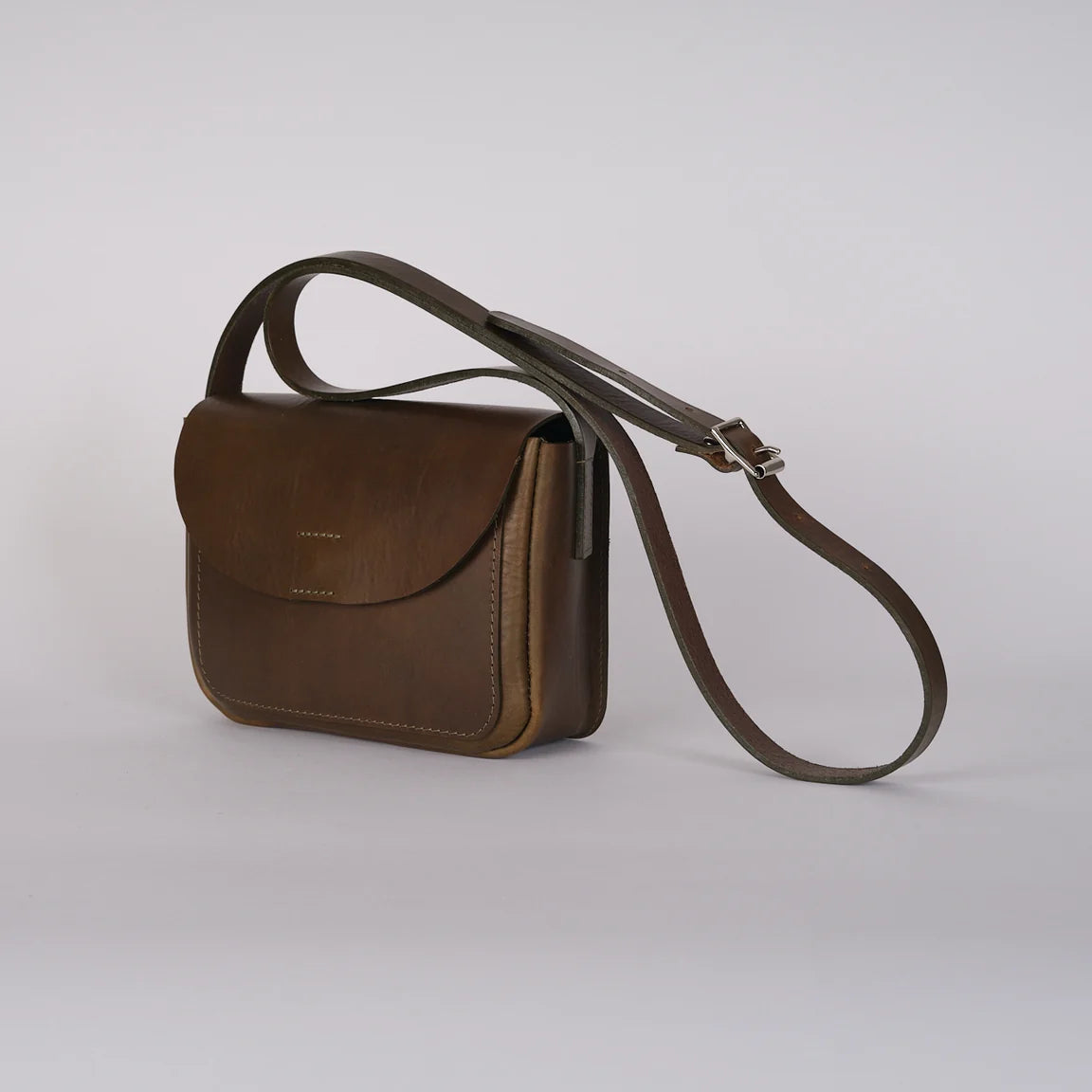 A sage green leather crossbody bag with a curved flap and a long adjustable strap, photographed on a light gray studio background.