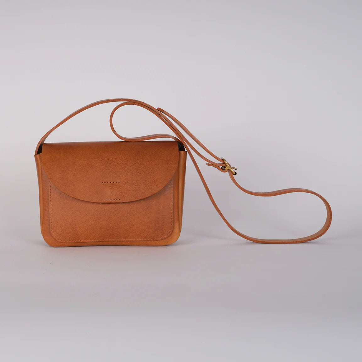 A small tan leather crossbody bag with a flap closure and a long adjustable strap, photographed on a clean light gray studio background.