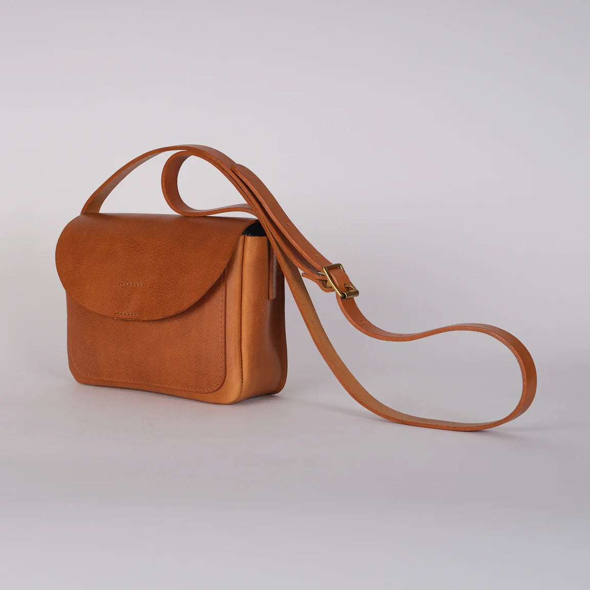 A small tan leather crossbody bag with a flap closure and a long adjustable strap, photographed on a clean light gray studio background, side view