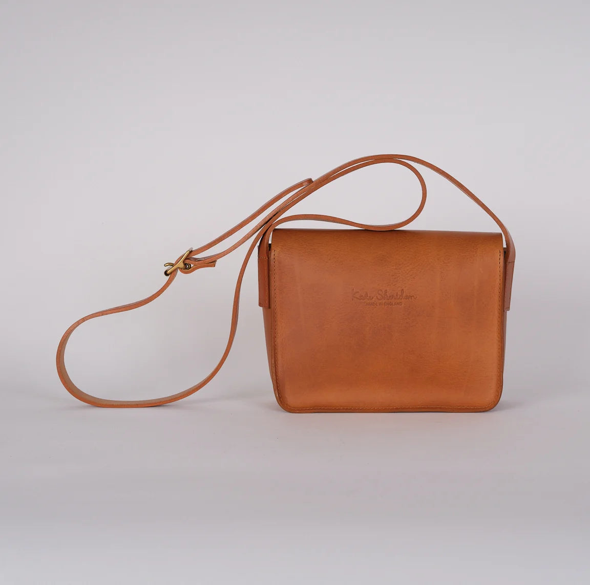 A small tan leather crossbody bag with a flap closure and a long adjustable strap, photographed on a clean light gray studio background, rear view.