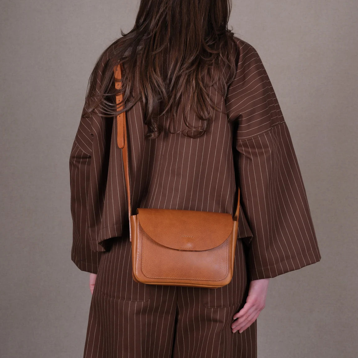 Women seen from behind wearing a brown striped coat with a small tan leather crossbody bag with a flap closure and a long adjustable strap, photographed on a clean light gray studio background.