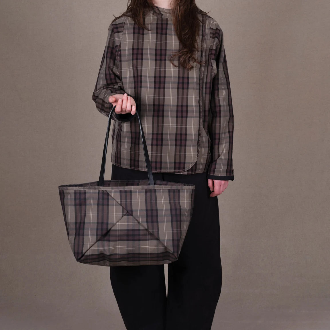 Person holding a large tote bag with a brown, gray, and black plaid pattern and black shoulder straps, shown against a plain white background.