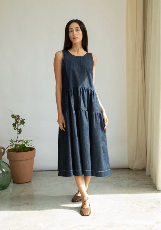 Woman wearing a dark blue dress standing in a minimalistic room with a plant and curtain.