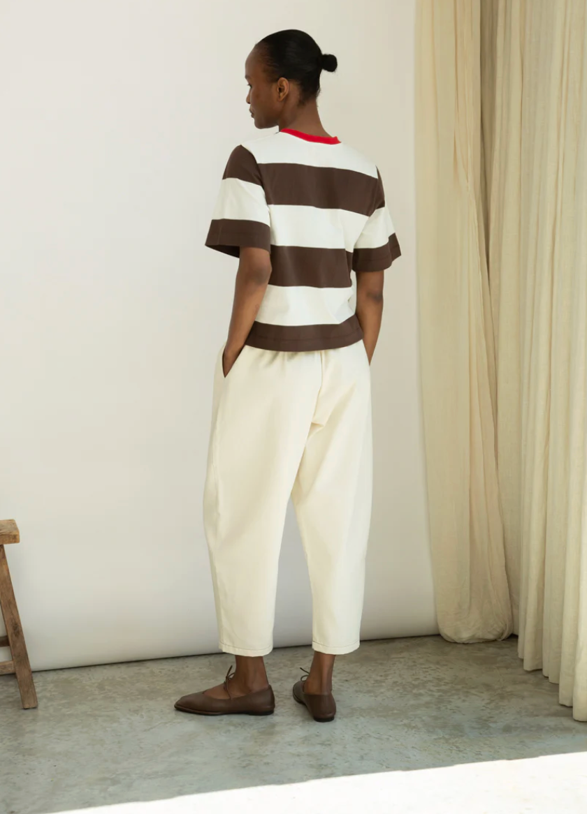 Person wearing a striped shirt and white pants standing against a neutral wall with a curtain.