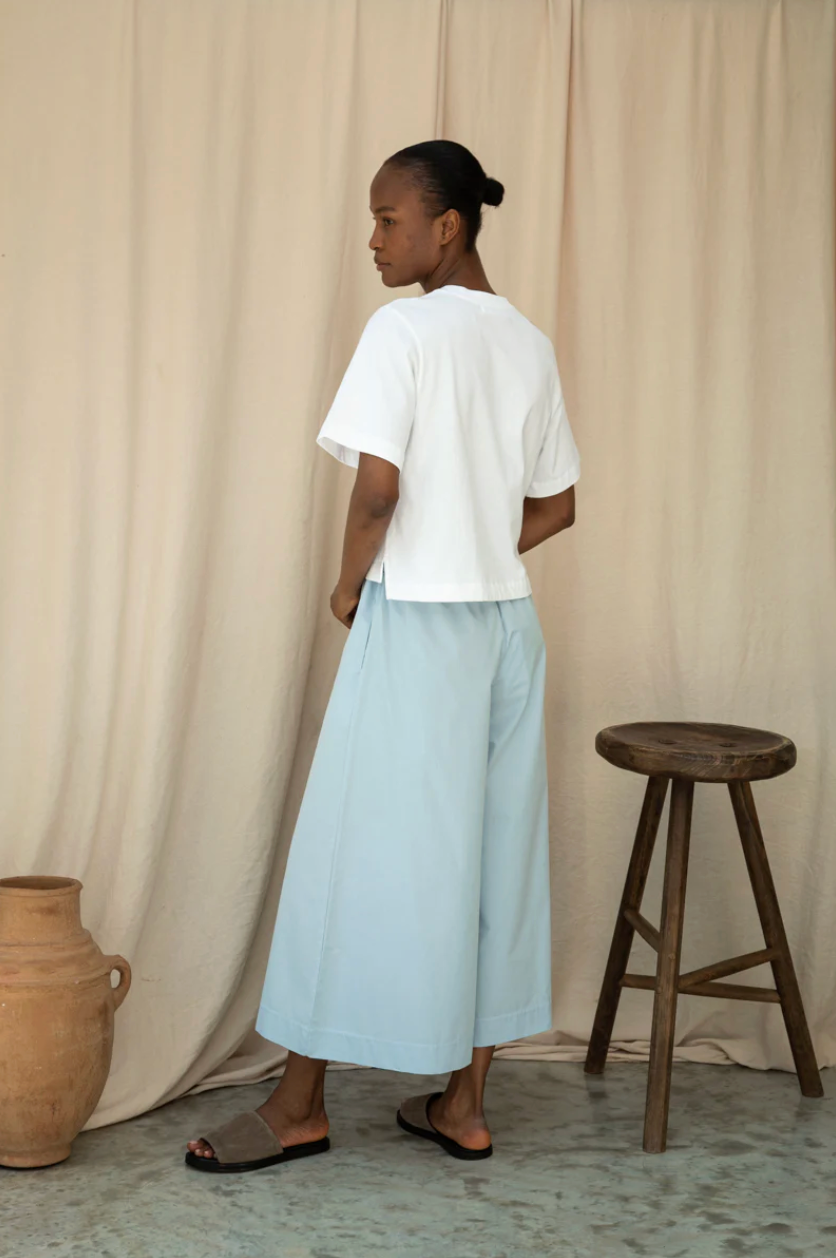 Woman wearing a white top and light blue pants standing in front of a beige curtain with a wooden stool and vase.