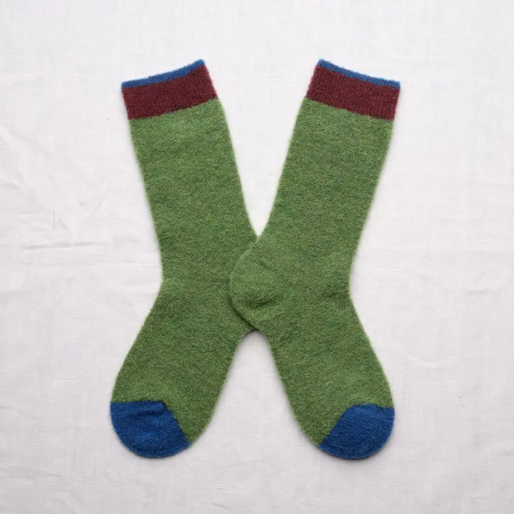 A pair of green knit socks with a dark red cuff stripe and blue toe caps, laid flat on a white fabric background.