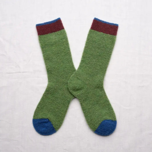 A pair of green knit socks with a dark red cuff stripe and blue toe caps, laid flat on a white fabric background.