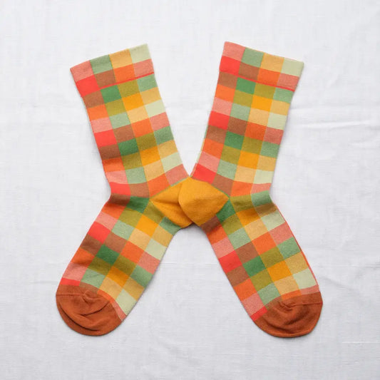A pair of gingham crew socks laid flat in an X shape on a white fabric background. The socks feature a multicolor pattern with mustard yellows, sage greens, and corals.