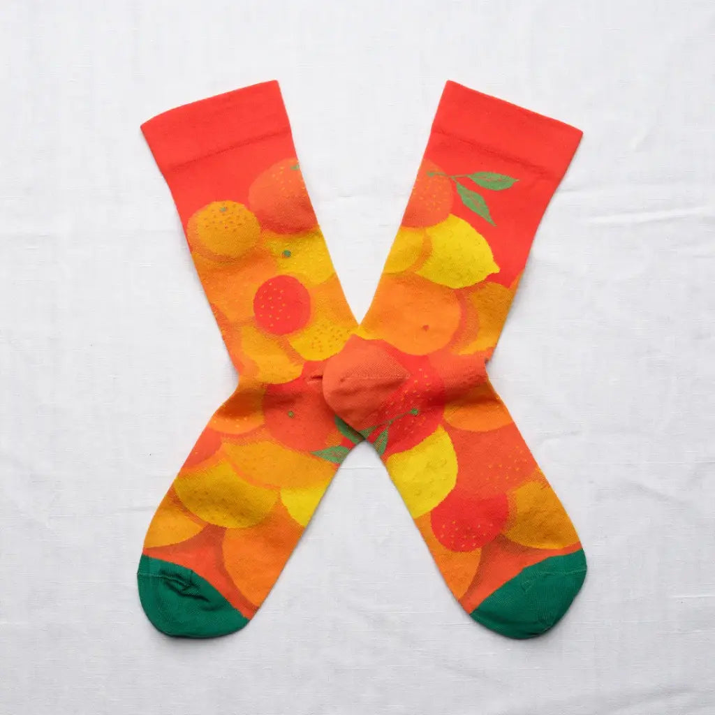 A pair of colorful crew socks in an X shape on a white background. The socks feature a group of citrus fruits on a vermillion background with green toes.