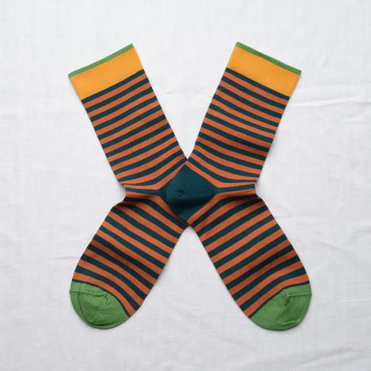 A pair of striped crew socks arranged in an X shape on a white fabric background. The socks feature horizontal stripes of dark teal and orange-brown, with a solid dark teal center, green toes, and yellow-green cuffs.