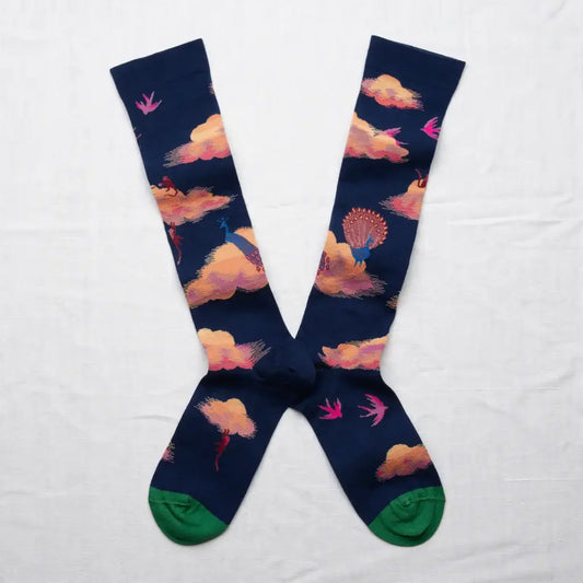 A pair of long crew socks in an X shape on a white background. The navy socks feature a dreamy cloud pattern with peacocks, birds, and little monkeys. The sock toe color is green.