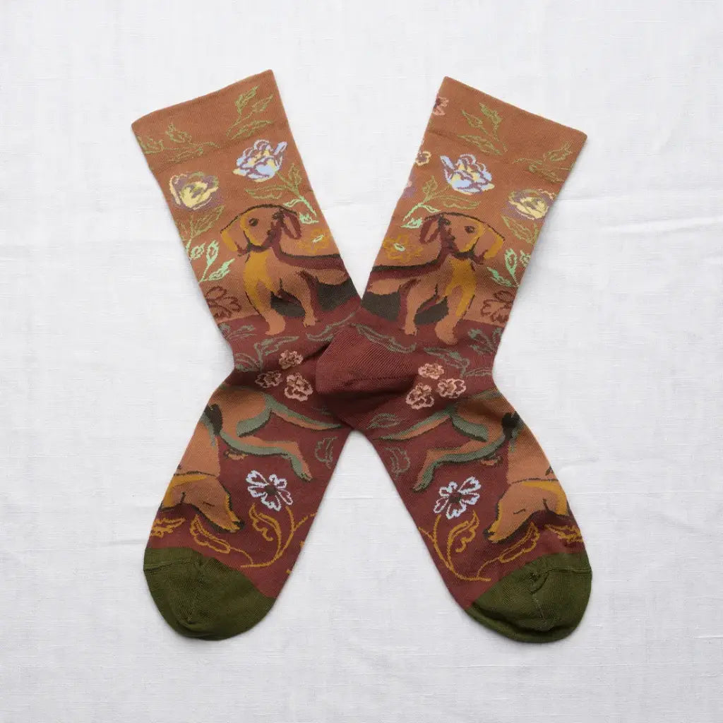 A pair of patterned crew socks laid flat on a white fabric background. The socks feature a warm brown and rust base with an all-over print of dachshunds, flowers, and green leaves, finished with solid olive green toes and heels.