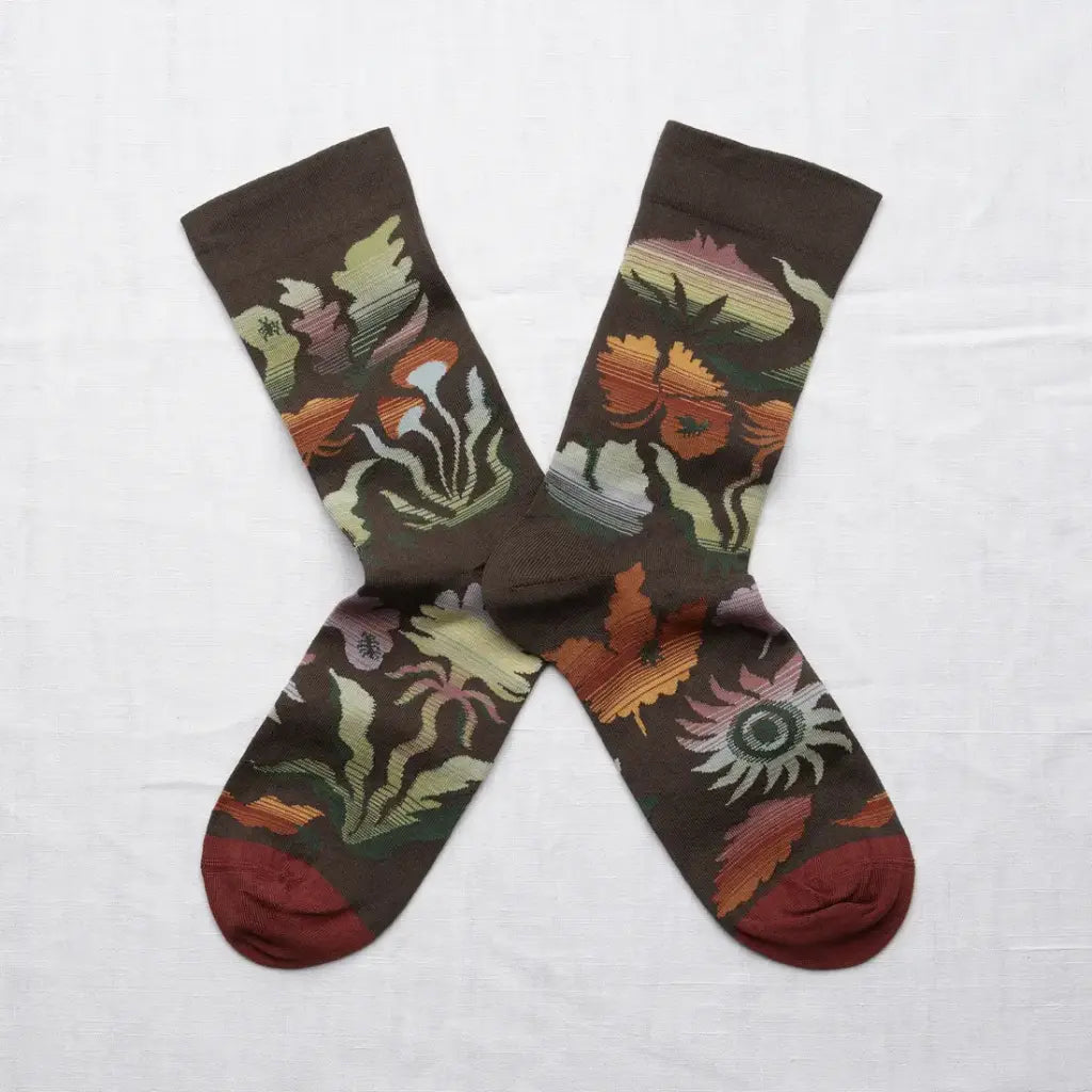 A pair of moss green crew socks in an X shape on a white background. The socks have a muted autumnal-rainbow of colors on a forest-inspired pattern. The sock toe color is maroon.