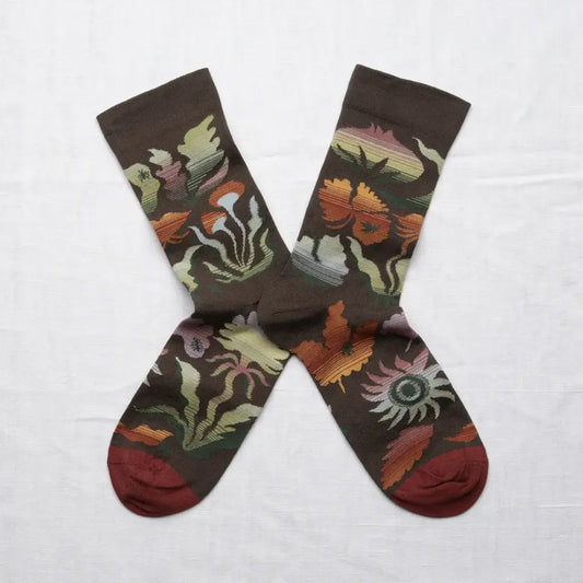 A pair of moss green crew socks in an X shape on a white background. The socks have a muted autumnal-rainbow of colors on a forest-inspired pattern. The sock toe color is maroon.