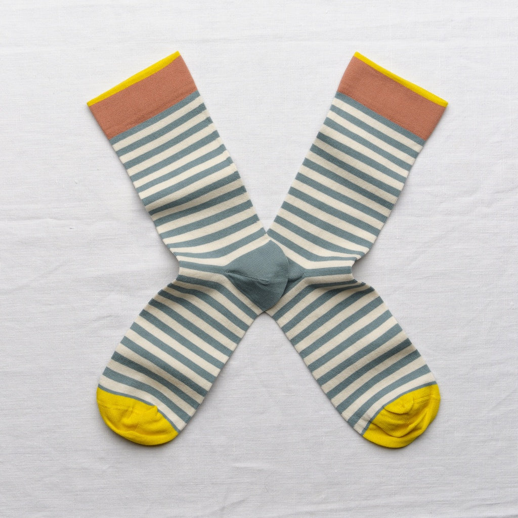 A pair of striped crew socks laid flat in an X shape on a white fabric background. The socks feature horizontal stripes in sage green and cream, with a solid sage green center, a muted terracotta cuff, and bright yellow toes and cuffs.