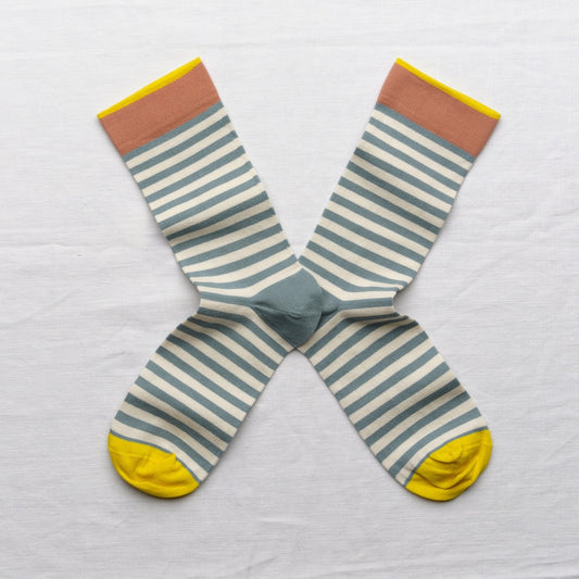 A pair of striped crew socks laid flat in an X shape on a white fabric background. The socks feature horizontal stripes in sage green and cream, with a solid sage green center, a muted terracotta cuff, and bright yellow toes and cuffs.