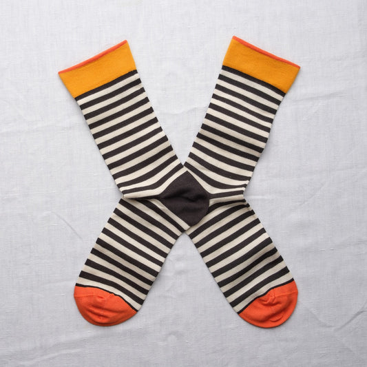 A pair of striped crew socks laid flat in an X shape on a white fabric background. The socks feature horizontal stripes in dark grey and cream, with a solid grey  center, a yellow and orange cuff, and bright orange toes.