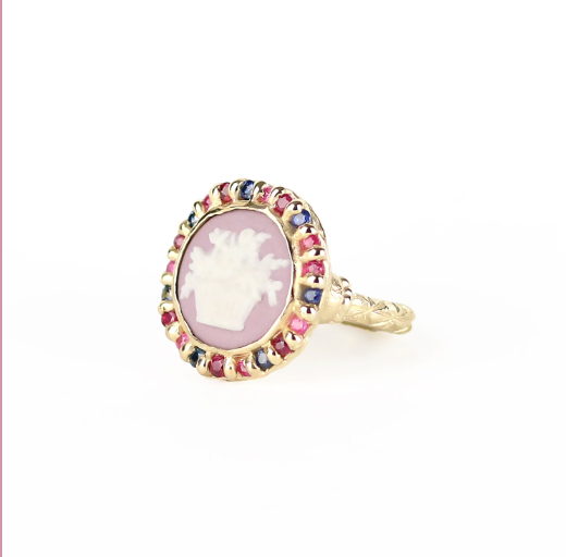 Side angle view of round gold ring with antique Wedgewood cameo garland motif on purple background surrounded by blue and pink sapphires and rubies