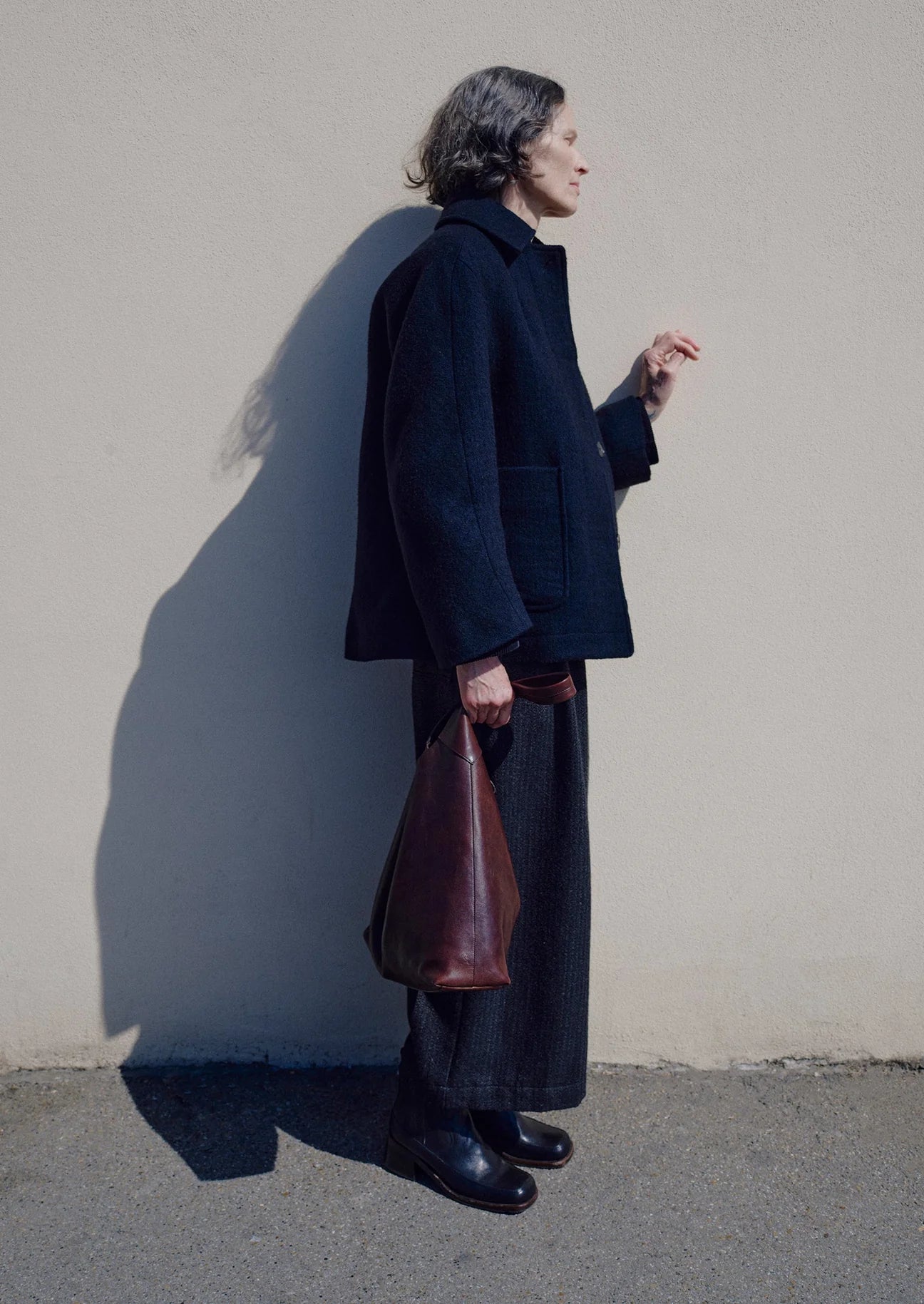 Person wearing a dark coat and carrying a brown leather bag against a plain wall.