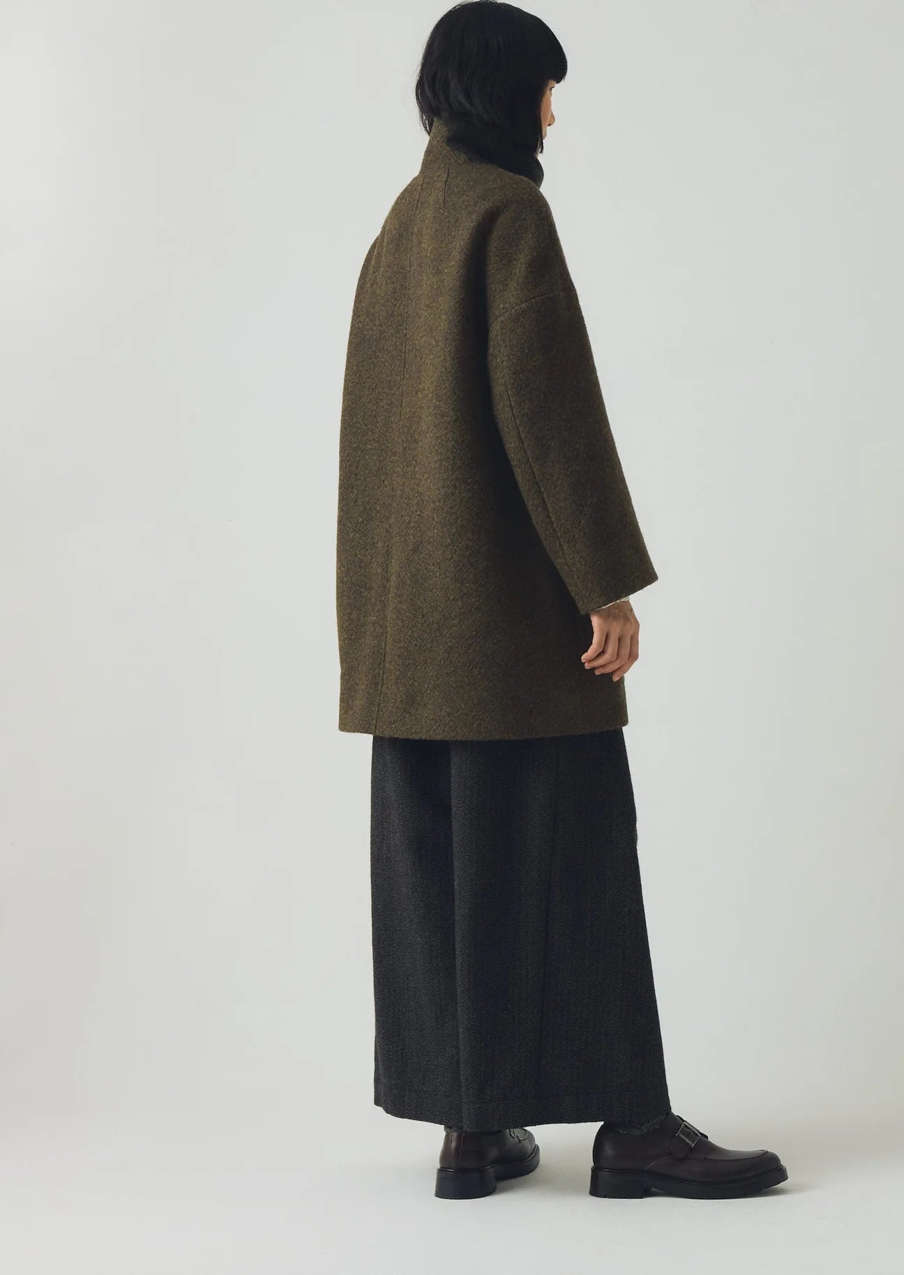 Person wearing a long olive green coat and dark pants on a white background