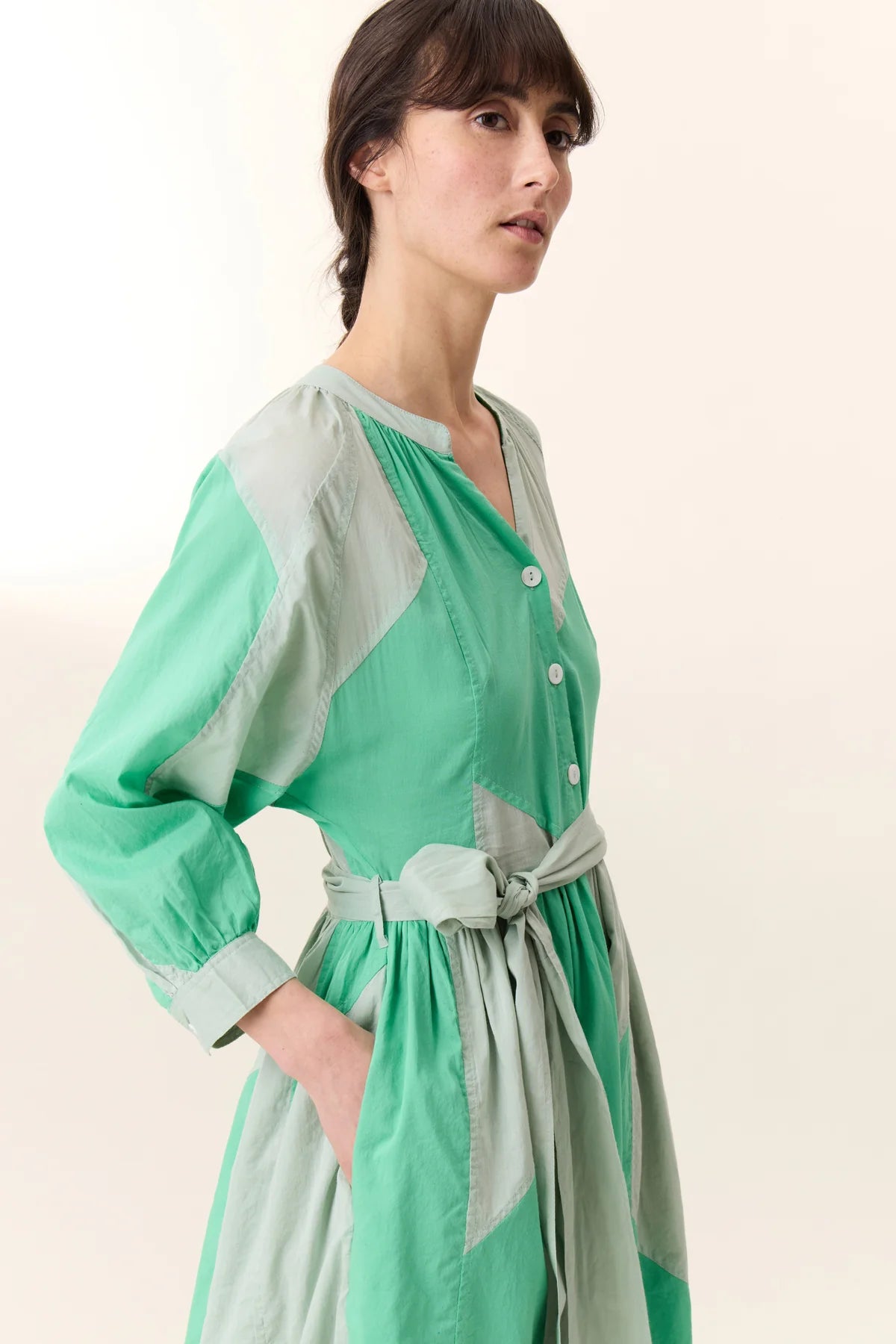 cropped photo of woman with long brown hair wearing green patch construction dress with tie belt. 