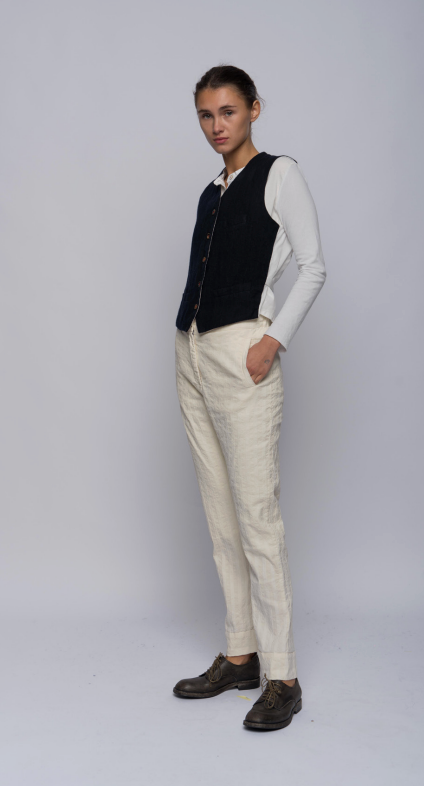 A model stands in profile wearing a dark navy vest over a white long-sleeve shirt, paired with off-white trousers and dark brown leather shoes, against a light gray studio background.