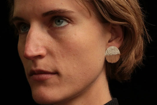 A close-up of a person's ear wearing a textured, circular brass earring against a solid black background.