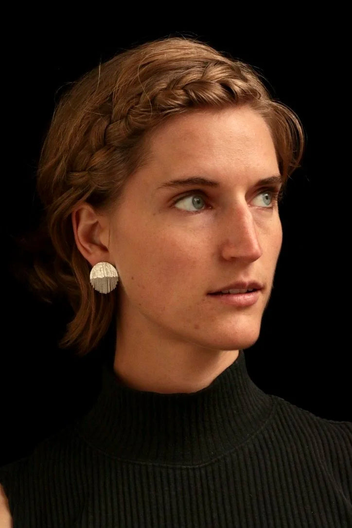 A close-up portrait of a woman with short brown hair wearing a textured silver disc earring, set against a solid black background.