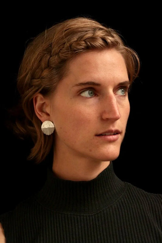 A close-up portrait of a woman with short brown hair wearing a textured silver disc earring, set against a solid black background.