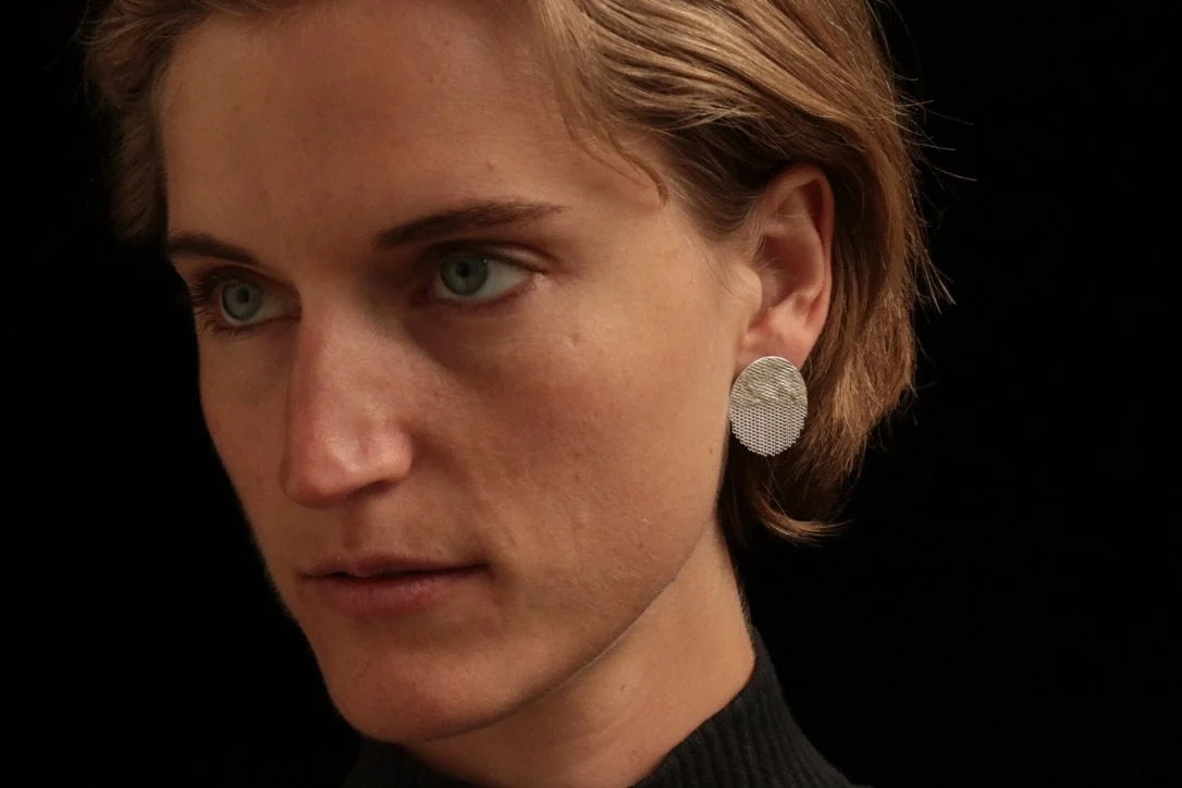 A close-up portrait of a woman with short light brown hair wearing a textured silver disc earring against a solid black background.
