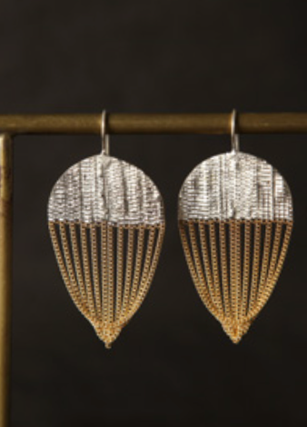 A pair of large teardrop-shaped dangle earrings with a textured silver top half and gold dangling chain-like fringe, hanging from a gold display stand against a dark background.