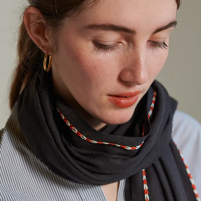 Graphite scarf with orange and cream stitch along border. Worn around neck and over left shoulder