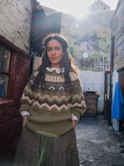 Person wearing a patterned sweater standing in an outdoor setting with a building and greenery in the background.