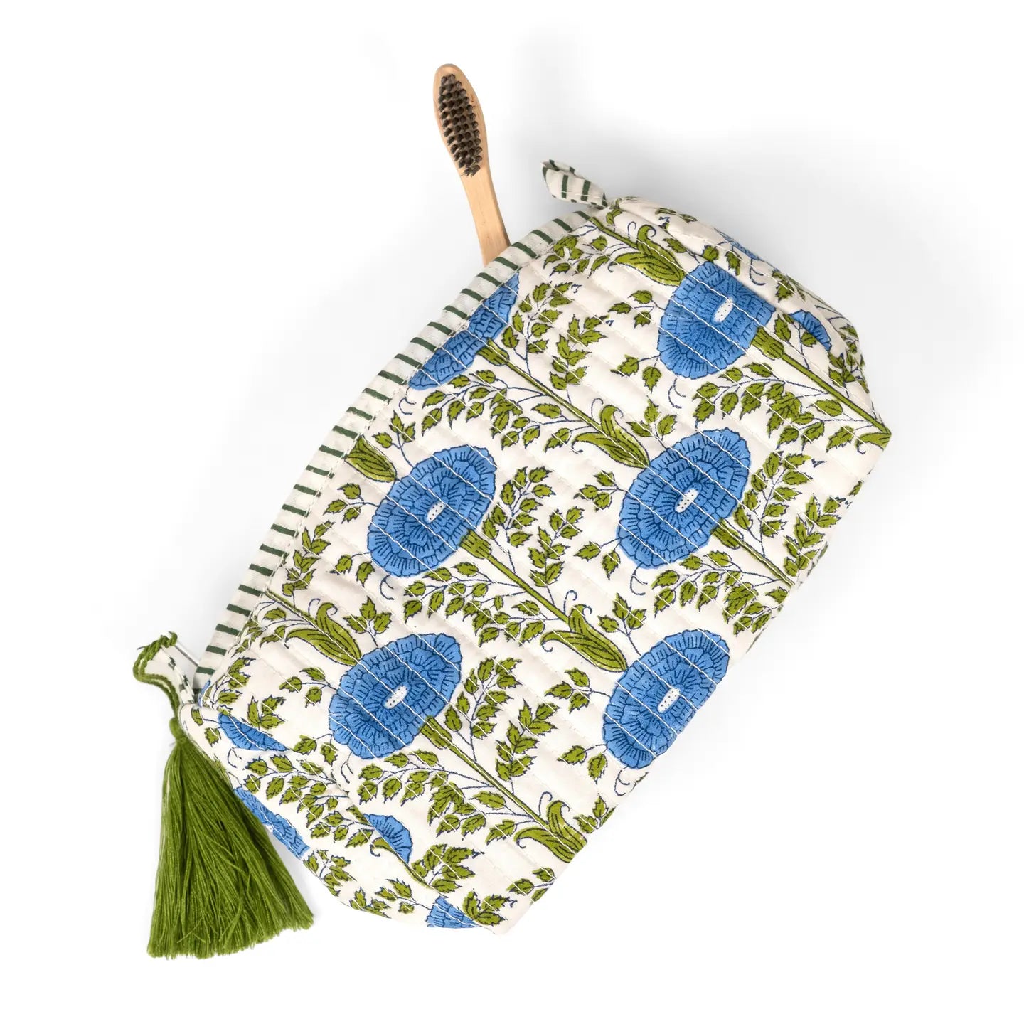 A fabric zippered pouch with a green and blue botanical block print on a cream background, featuring a green tassel and a wooden toothbrush tucked inside, shot on a white background.