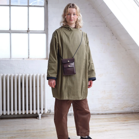 A person wearing an olive green hooded jacket and brown pinstriped trousers, standing in a bright attic room with white brick walls, a large skylight, and a radiator.