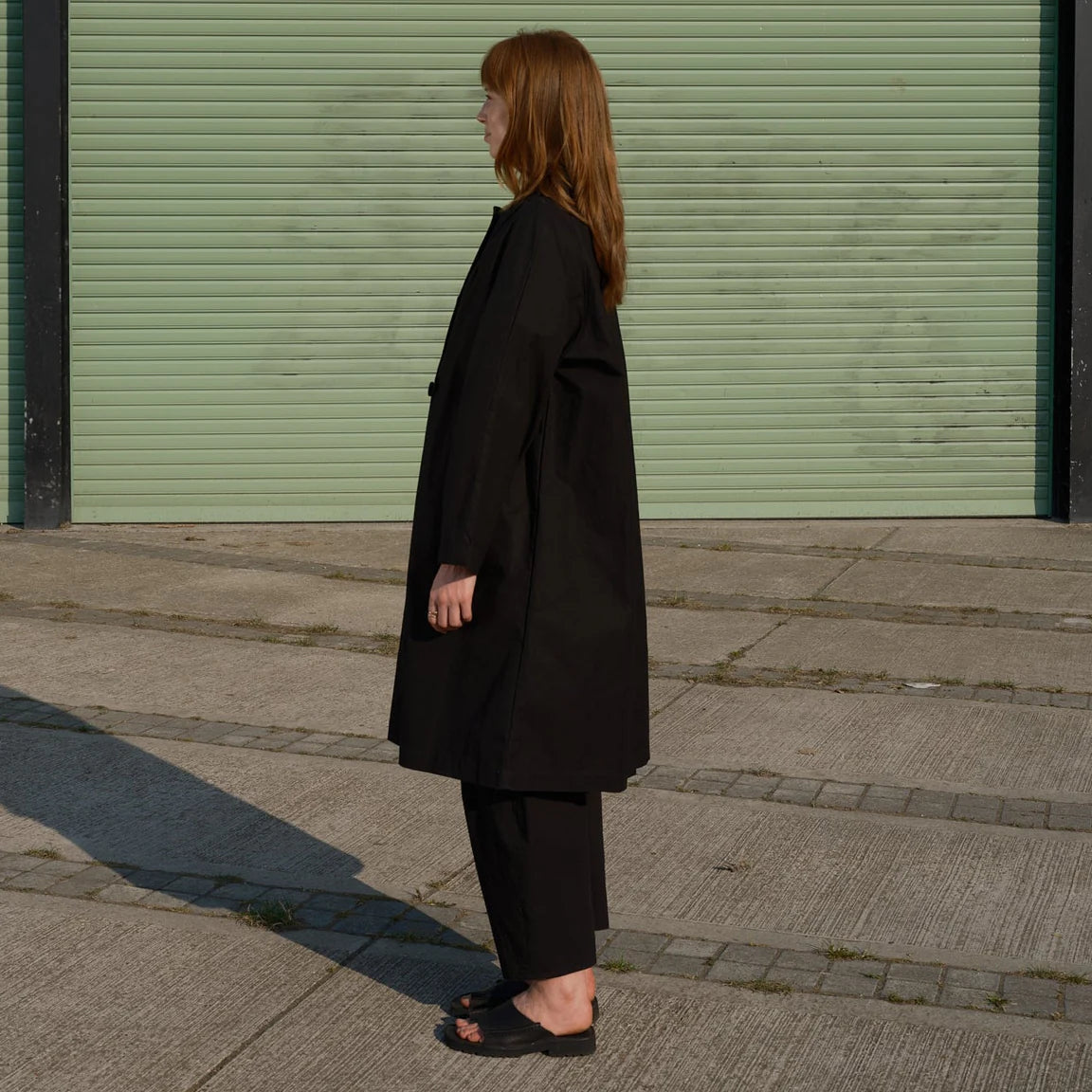 Person wearing a black coat standing on a sidewalk with a green shutter in the background