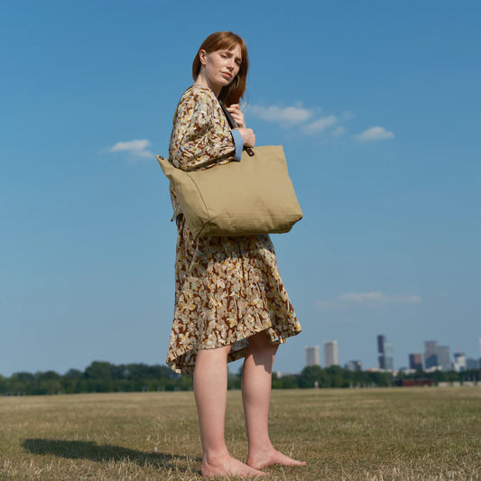 Woman holding a beige tote bag in an open field with a city skyline in the background