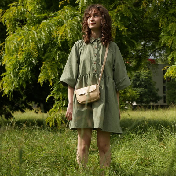 Picture,  of woman with long wavy brown hair in green dress wearing a light brown handbag. Standing in a field of grass in front of a tree.