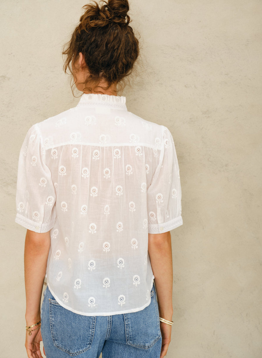 Picture of young women with her back to camera. With curly brown hair in a bun and brown eyes. Wearing a white short sleeve shirt and denim pants. Against a stone colored background.