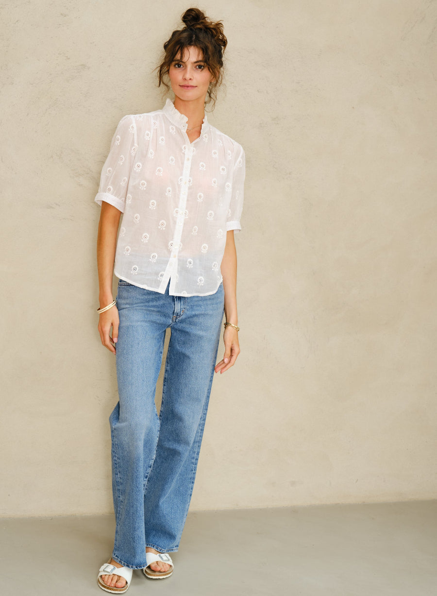 Picture of young women, full length. With curly brown hair in a bun and brown eyes. Wearing a white short sleeve shirt and denim pants. Against a stone colored background.