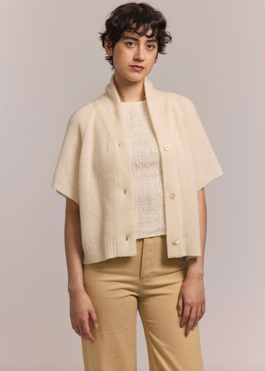 Woman with short wavy dark brown hair wearing an ivory short sleeve cashmere cardigan with a silk blouse and tan colored pants