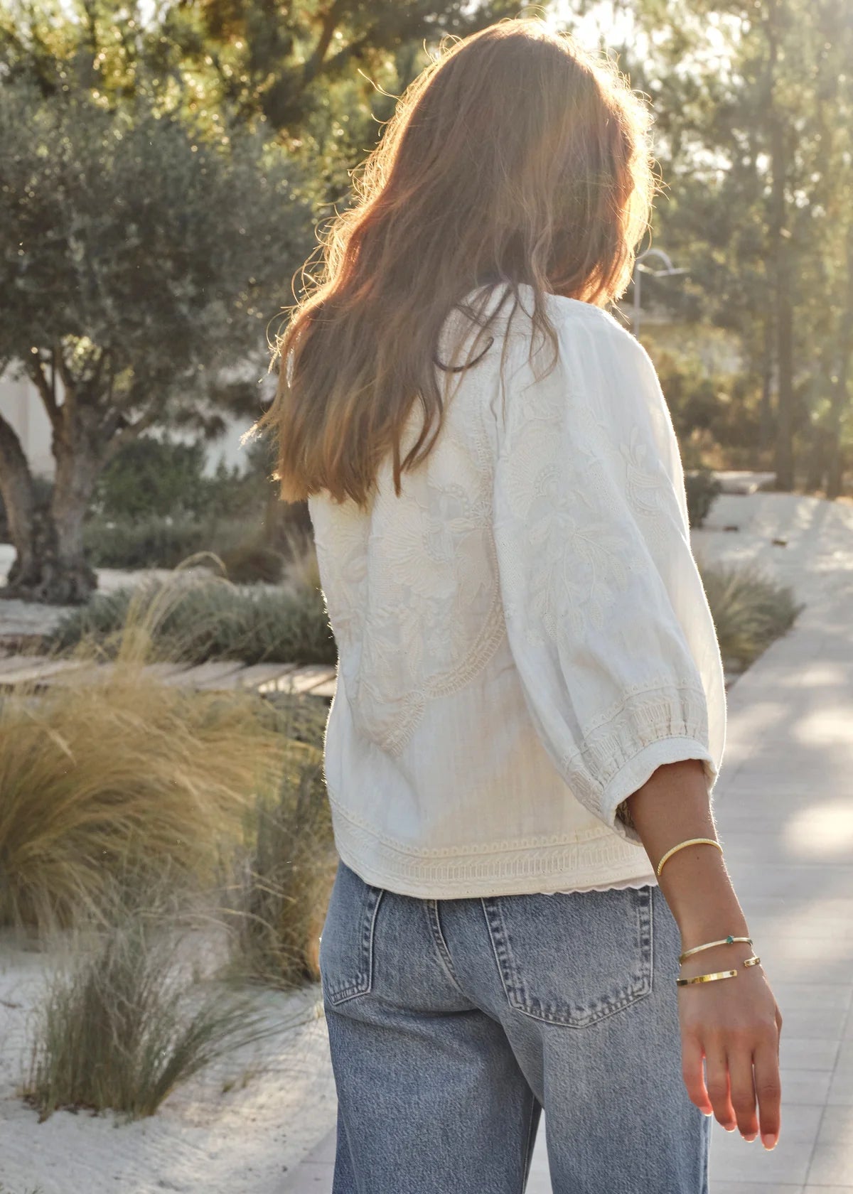 Person wearing a light-colored shirt and blue jeans walking outdoors with trees and plants in the background