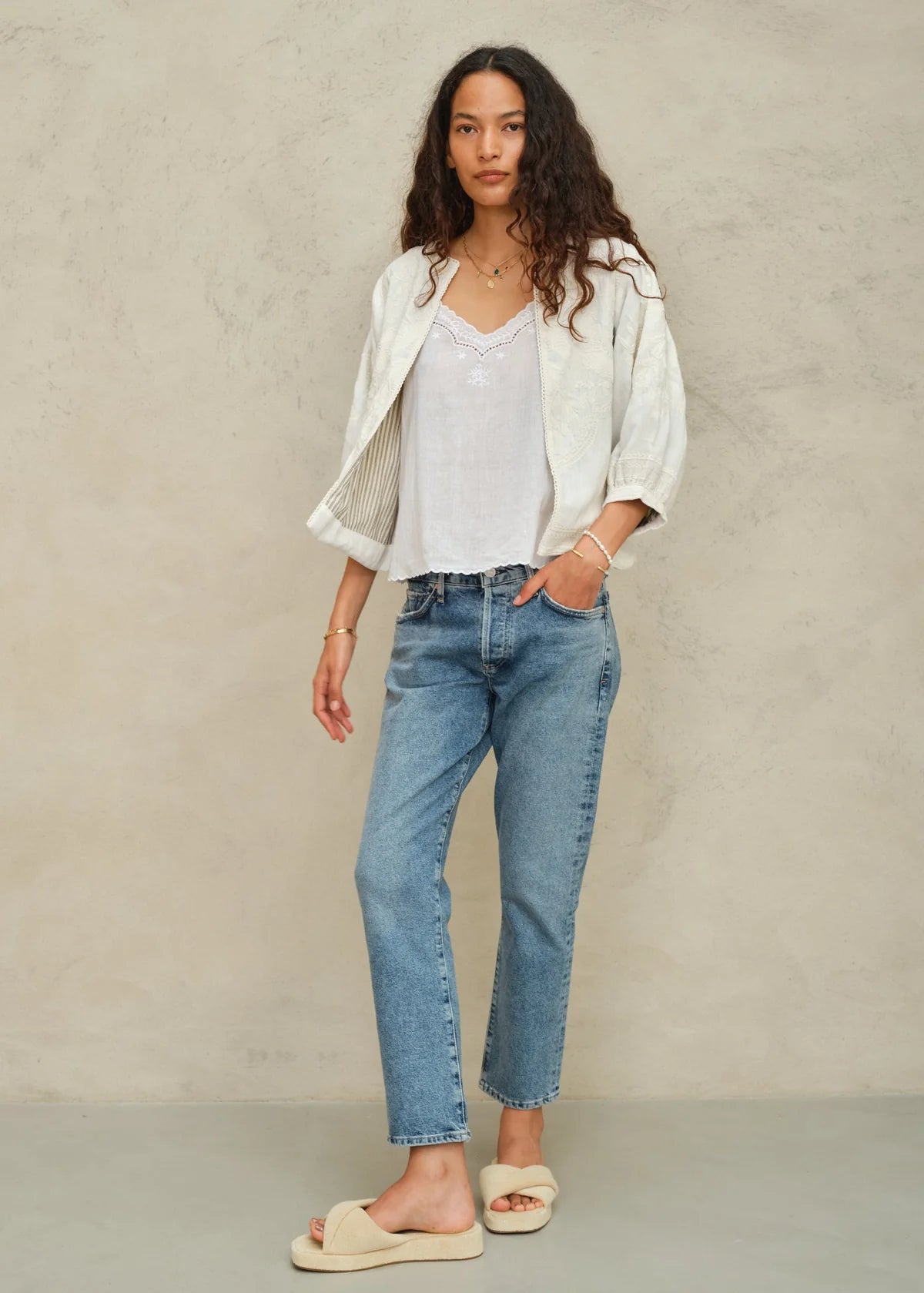 Woman wearing a white jacket, white shirt, blue jeans, and beige sandals against a neutral background